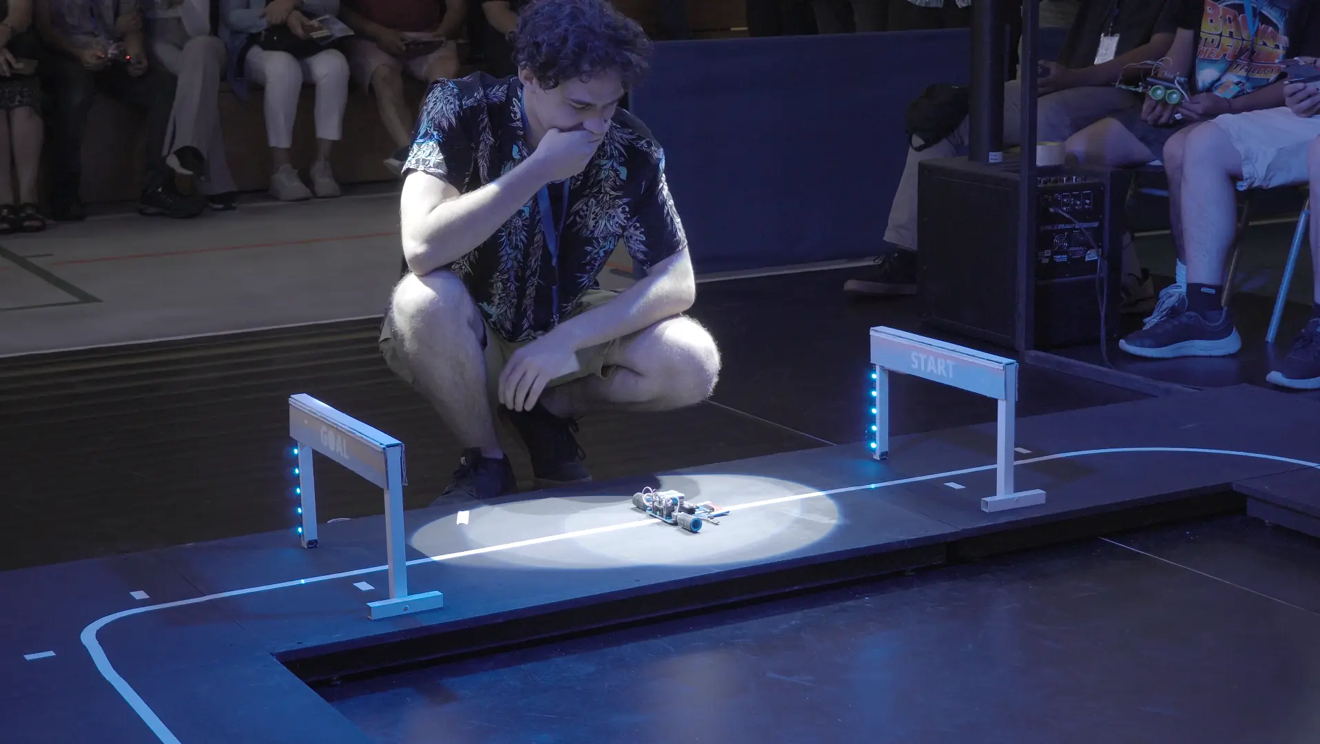 Un competidor mira su robot en la partida de la pista de Robotracer, entre los arcos de meta y de inicio, frente al público